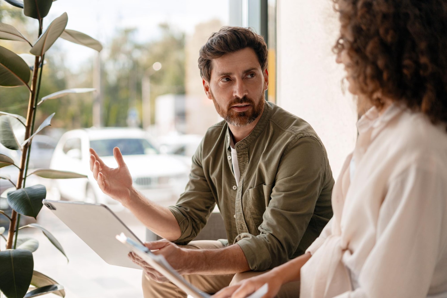 A man and a woman sitting by a window talking with a laptop between them